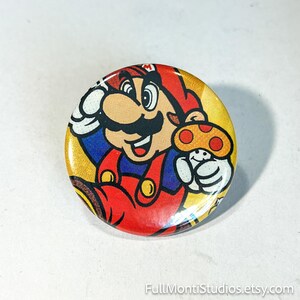 May include: A round button featuring a colorful illustration of the video game character Mario holding a mushroom. Mario is depicted in his signature red hat and overalls, with a yellow background.