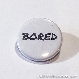 May include: A round, white button with the word "BORED" in black, handwritten-style lettering. The button has a glossy finish and a slight shadow, suggesting it is raised from the surface. The button is a simple, yet expressive accessory.