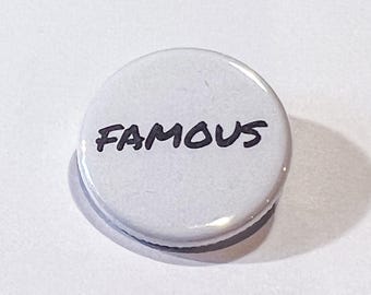 May include: A white button with the word "FAMOUS" in black handwritten font. The button is round and has a glossy finish. The button is a small accessory.