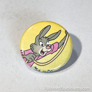 May include: A round button with a cartoon rabbit illustration. The rabbit is gray with a pink shirt and is set against a yellow background. The button has a glossy finish. The Etsy shop name "FullMontiStudios.etsy.com" is printed at the bottom.