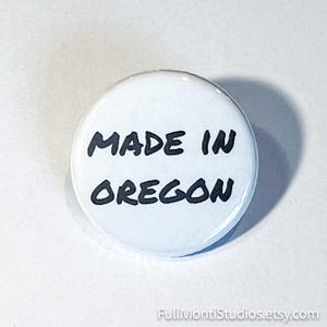 May include: A white button with the text "MADE IN OREGON" in black, handwritten-style lettering. The button is round and has a glossy finish. The background is a light, neutral color.