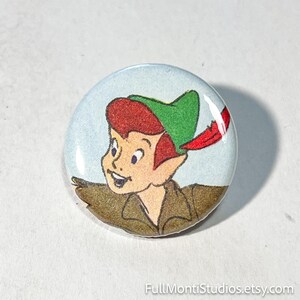 May include: A round button featuring a cartoon illustration of Peter Pan. The character has red hair, a green hat with a red feather, and a brown jacket. The button has a light blue background.