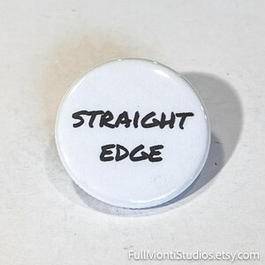 May include: A white button with the words "STRAIGHT EDGE" in black, handwritten-style lettering. The button is round and has a glossy finish. The button is a simple, bold statement.