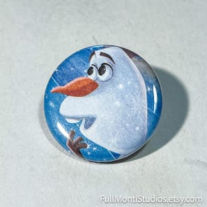 May include: A round button featuring Olaf from Disney's Frozen. The button has a blue background with white stars and a smiling Olaf with an orange carrot nose. The text "FullMontiStudios.etsy.com" is at the bottom.