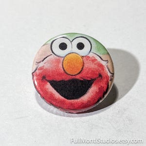 May include: A round button featuring a watercolor illustration of a smiling, red character with a large orange nose, black mouth, and white eyes. The button has a green and tan background. The Etsy shop name "FullMontiStudios.etsy.com" is printed at the bottom.
