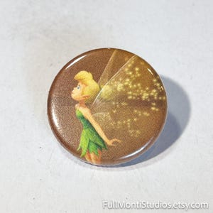 May include: A round button featuring a cartoon fairy with blonde hair and green dress. The fairy has translucent wings with gold glitter accents. The button has a brown background and a glossy finish. The Etsy shop name "FullMontiStudios.etsy.com" is at the bottom.