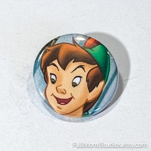 May include: A round button featuring a cartoon illustration of a boy with brown hair, a green hat, and a smiling expression. The button has a glossy finish and a light blue background. The button is a collectible item.
