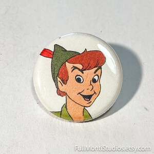May include: A round button featuring a cartoon illustration of Peter Pan. The character has red hair, a green hat with a red feather, and a green shirt. The button has a glossy finish and a white background.