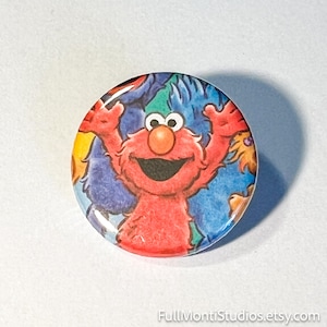 May include: A round button featuring the Sesame Street character Elmo with his arms raised. The button has a colorful background of blue, green, and yellow. The button is a collectible item.