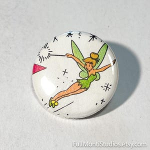 May include: A round, white button featuring a cartoon illustration of a fairy with green wings and a green dress. The fairy is holding a wand and surrounded by stars and other decorative elements.