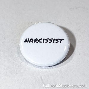 May include: A white button with the word "NARCISSIST" in black, handwritten-style lettering. The button is round and has a glossy finish. The background is a light, textured surface.