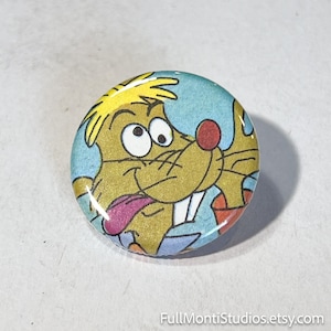 May include: A round button featuring a cartoon character with yellow fur, a red nose, and a pink tongue. The character is set against a light blue background. The button has a glossy finish.