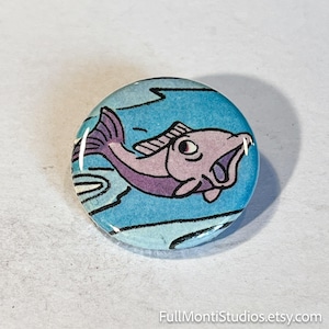 May include: A round button with a cartoon illustration of a purple fish with its mouth open, set against a blue background with water lines. The button has a glossy finish. The text "FullMontiStudios.etsy.com" is at the bottom.