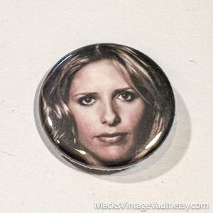 May include: A round button featuring a portrait of a woman with blonde hair and a serious expression. The button has a glossy finish and a dark border. The Etsy shop name "MacksVintageVault.etsy.com" is printed at the bottom.