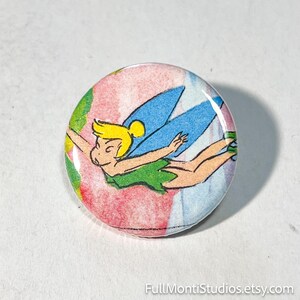 May include: A round button featuring a cartoon illustration of a fairy with blonde hair, blue wings, and a green dress. The background has a colorful, abstract pattern. The button is a collectible item.