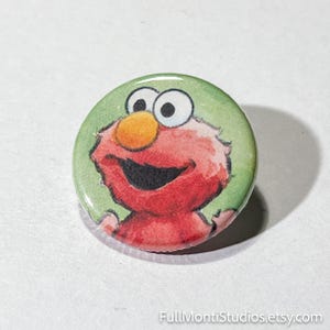 May include: A round button featuring a watercolor illustration of Elmo from Sesame Street. The button has a green background and a glossy finish. Elmo's face is red with a large orange nose, black eyes, and a smiling mouth. The text "FullMontiStudios.etsy.com" is at the bottom.