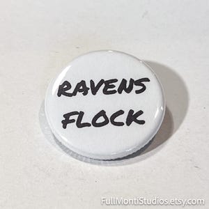 May include: A white button with the words "RAVENS FLOCK" in black, bold lettering. The button has a glossy finish and a slightly raised edge. The text is centered on the button.