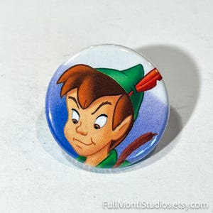 May include: A round button featuring a cartoon illustration of a boy with brown hair, wearing a green hat with a red feather. The button has a blue and white background and the text "FullMontiStudios.etsy.com".