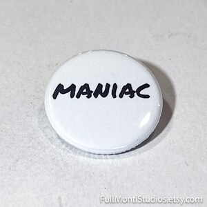 May include: A white button with the word "MANIAC" in black, handwritten-style lettering. The button is round and has a glossy finish. The background is a light gray surface.