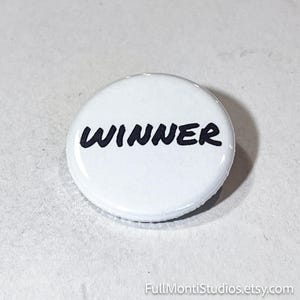 May include: A white, round button with the word "WINNER" in bold, black letters. The button has a glossy finish and is likely a pin or badge. The background is a light gray surface.