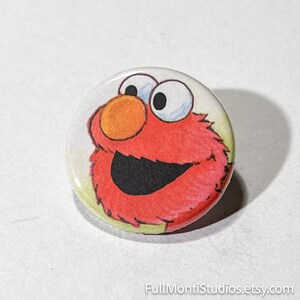 May include: A round button featuring a colorful illustration of Elmo from Sesame Street. The button has a white background with Elmo's red fur, orange nose, black mouth, and large white eyes. The Etsy shop name, FullMontiStudios.etsy.com, is printed at the bottom.