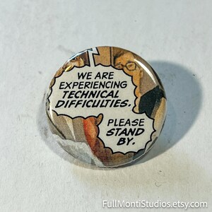 May include: A round button with a comic book style design. The button features a speech bubble with the text "WE ARE EXPERIENCING TECHNICAL DIFFICULTIES. PLEASE STAND BY."