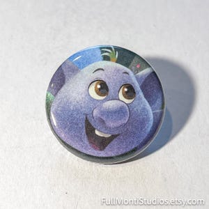 May include: A round button featuring a cartoon character with a purple face, large brown eyes, and a wide smile. The character has large ears and a small tuft of green hair. The button has a glossy finish and a black border.