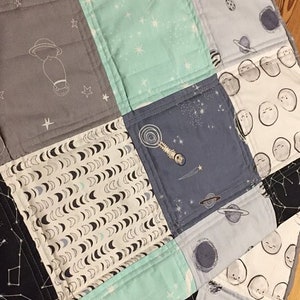 May include: A patchwork quilt with a space theme. The quilt is made of various fabrics with different patterns, including stars, moons, planets, and rockets. The quilt is in shades of blue, gray, and white.