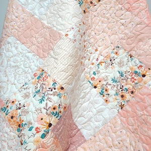 May include: A quilted blanket with a patchwork design. The quilt features various floral and geometric patterns in shades of peach, white, and cream. The stitching creates a textured effect, enhancing the overall aesthetic of the handmade quilt.