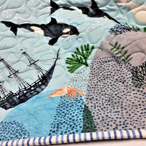 May include: A quilted fabric featuring an underwater scene with orcas, a sailing ship, and various marine life. The design includes blue, gray, and orange elements, with a striped border.
