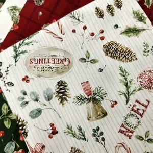 Pode incluir: Um tecido de cor creme com uma estampa natalina de pinhas, folhas de azevinho, bagas vermelhas, sinos e bengalas doces. O texto diz Season's Greetings, Noel e North Pole. Camadas adicionais de tecido em vermelho e verde escuro s&atilde;o vis&iacute;veis.