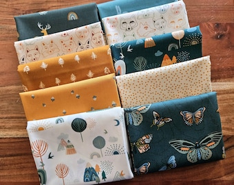 May include: A collection of folded fabric pieces with various patterns. Designs include foxes, deer, trees, butterflies, and mountain scenes. Colors include teal, mustard yellow, cream, and white. The fabrics are arranged on a wooden surface.