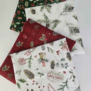 Pode incluir: Quatro quadrados de tecido dobrados com padr&otilde;es natal&iacute;cios. Um &eacute; verde escuro com bagas vermelhas, um branco com ramos e pinhas, um vermelho com padr&atilde;o xadrez e azevinho, e um branco com pinhas, bengalas doces e texto.