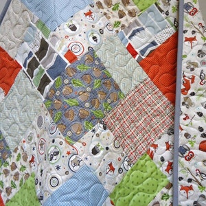May include: A patchwork quilt with a woodland theme. The quilt features squares of various fabrics with patterns of foxes, acorns, and geometric designs in shades of orange, green, blue, and gray. The quilt is bordered with a light gray fabric.