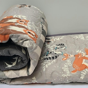 May include: A gray and white quilted blanket with a woodland animal print. The blanket features deer, raccoons, and the text "Hello, Good".