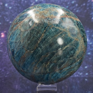 May include: A teal and brown gemstone sphere, polished to a high sheen, presented on a clear stand. The surface of the orb displays natural veining. The backdrop is a starry, cosmic design.