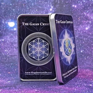 Może przedstawiać: Dwa srebrne pudełka z czarnymi i niebieskimi wzorami. Pudełko po lewej stronie ma wz&oacute;r geometryczny i tekst "THE GAIAN CRYSTALS" oraz "www.thegaiancrystals.com". Pudełko po prawej stronie ma globus z geometrycznym wzorem i tekst "THE GAIAN CRYSTALS" oraz "ORACLE AFFIRMATION DECK 78 CARDS AND GUIDE BOOKLET".