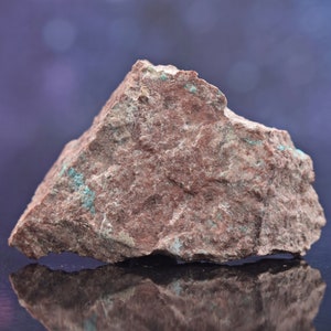 Może przedstawiać: Szorstki, brązowy kamień z zielonymi wrostkami mineralnymi. Kamień ma naturalną, nierówną powierzchnię i jest oświetlony miękkim, rozproszonym światłem.