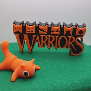 May include: A bright orange cat figurine in a playful pose with a raised tail, positioned in front of a black and orange sign that features the word WARRIORS in large letters topped with six distinct square icons representing various symbols.