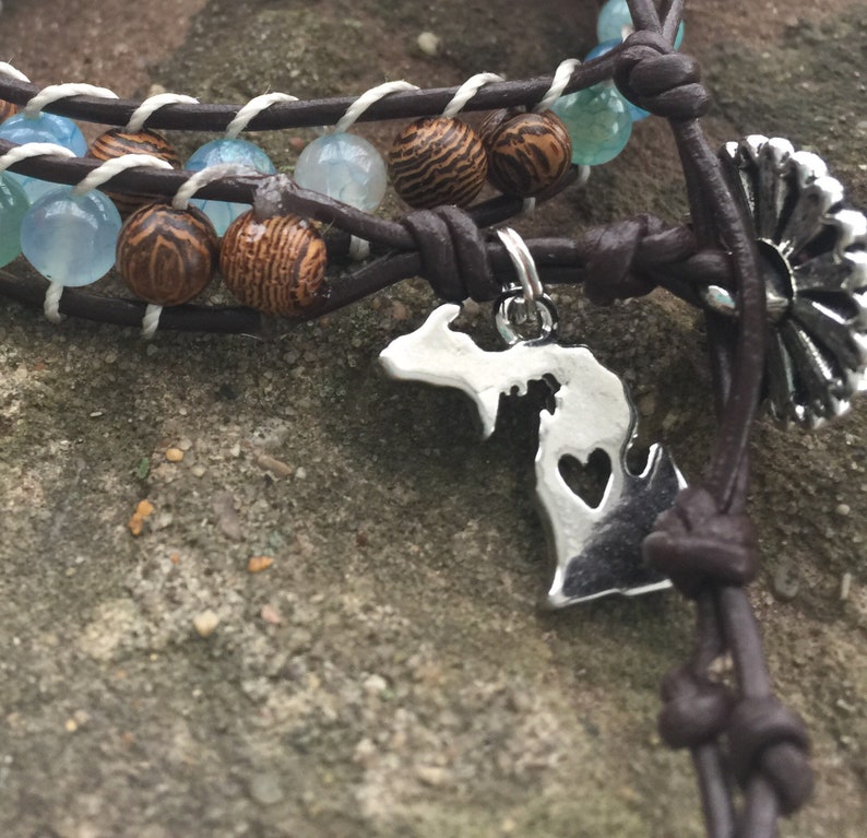Op de afbeelding: Een bruin leren armband met blauwe en bruine kralen en een zilveren bedel in de vorm van de staat Michigan met een uitgesneden hart.