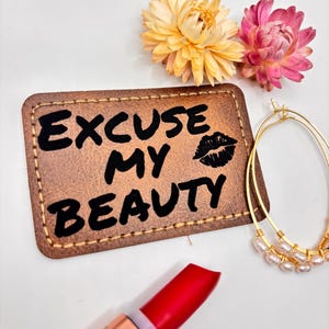 Puede incluir: Un parche de cuero marrón con el texto "EXCUSE MY BEAUTY" y una huella de labios negra. Un lápiz labial rojo, pendientes de aro dorados con perlas y dos flores secas también están en la imagen.