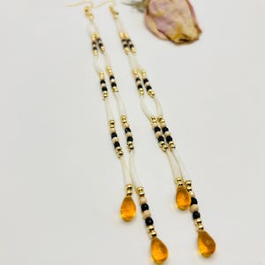 May include: A pair of long dangle earrings featuring white hair pipe beads, small black beads, and metallic gold-tone spacer beads. Each earring terminates in a translucent amber-colored teardrop-shaped pendant suspended from a gold-tone hook.