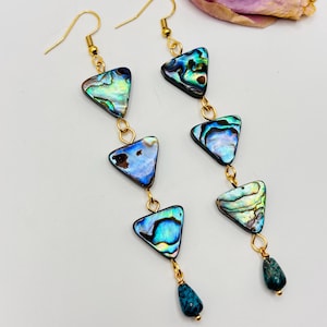Può includere: Un paio di lunghi orecchini pendenti con tre pezzi triangolari di conchiglia di abalone dai toni iridescenti blu e verdi, collegati da anellini in metallo dorato e rifiniti con una piccola pietra blu scuro a forma di goccia.