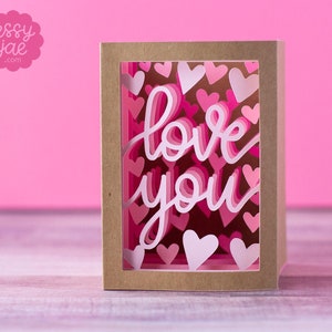 Puede incluir: Una tarjeta marr&oacute;n con una ventana recortada. La ventana est&aacute; llena de corazones de papel rosa y blanco y las palabras "love you" en papel rosa y blanco.