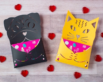 May include: Two paper gift card holders, one black pug and one yellow cat, each with a gift card inside. Red heart-shaped decorations are scattered around the holders on a light wood surface. The pug and cat have cut-out facial features.