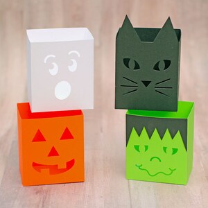 Pode incluir: Quatro caixas de papel com tema de Halloween com rostos recortados. As caixas estão empilhadas em dois pares. A caixa superior é branca com um rosto de fantasma, a segunda caixa é preta com um rosto de gato, a terceira caixa é verde com um rosto de Frankenstein e a caixa inferior é laranja com um rosto de abóbora.