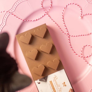 May include: A brown chocolate mold with six heart-shaped cavities. The mold is on a pink plate with a red and white string tied in a heart shape.