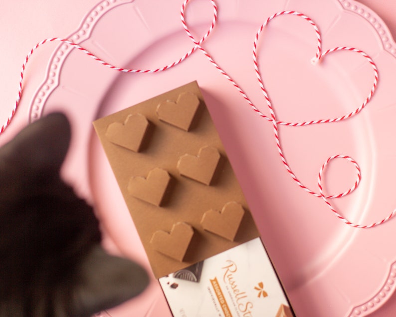 May include: A brown chocolate mold with six heart-shaped cavities. The mold is on a pink plate with a red and white string tied in a heart shape.