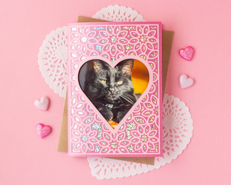 May include: Pink card with a heart-shaped cutout, featuring a black cat inside the cutout. The card has a decorative design of pink and white flowers.