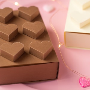 May include: A brown box with six heart-shaped compartments, perfect for holding small gifts or treats. The box is sitting on a pink surface with string lights and pink hearts.